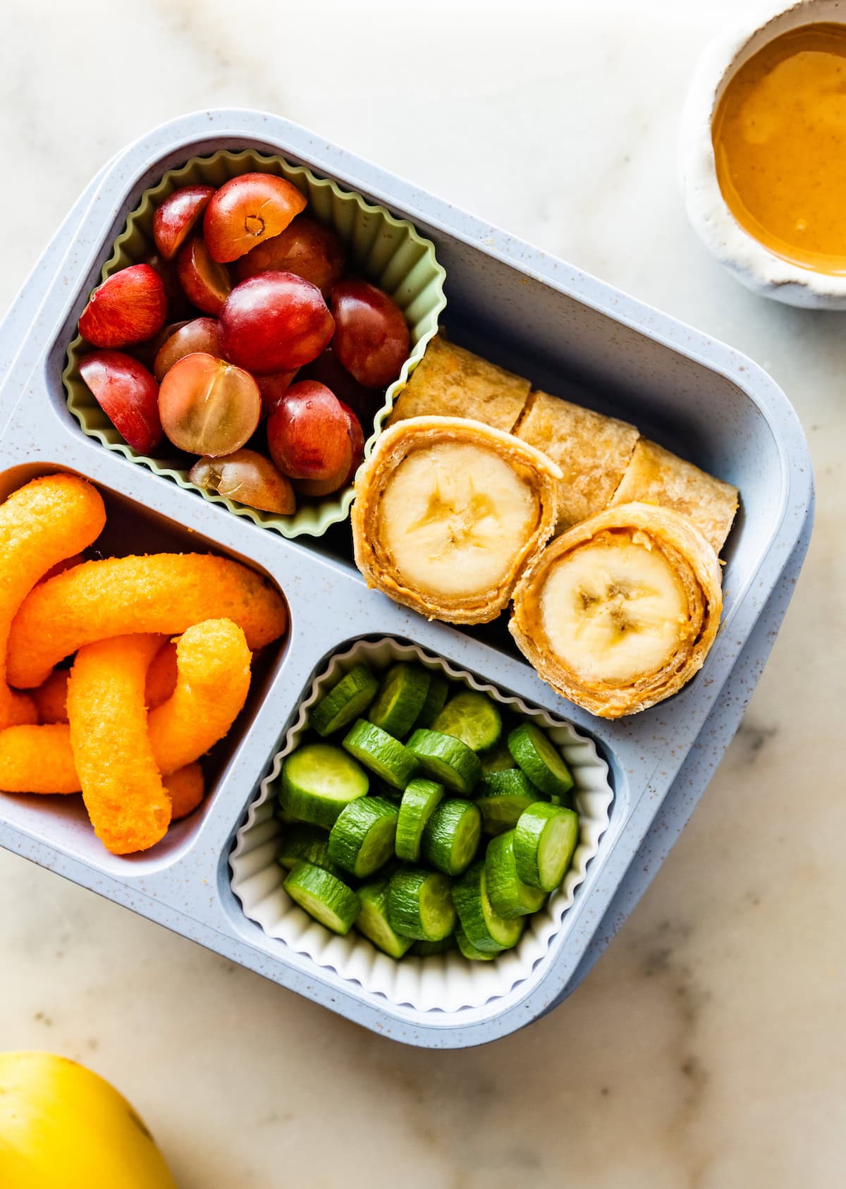 A lunchbox filled with banana sushi roll ups, grapes, cheetos, and cucumbers.