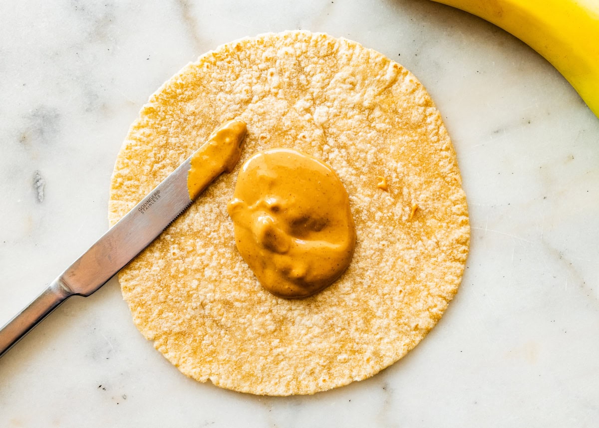 A nut butter is about to be spread on a whole wheat tortilla with a butter knife.