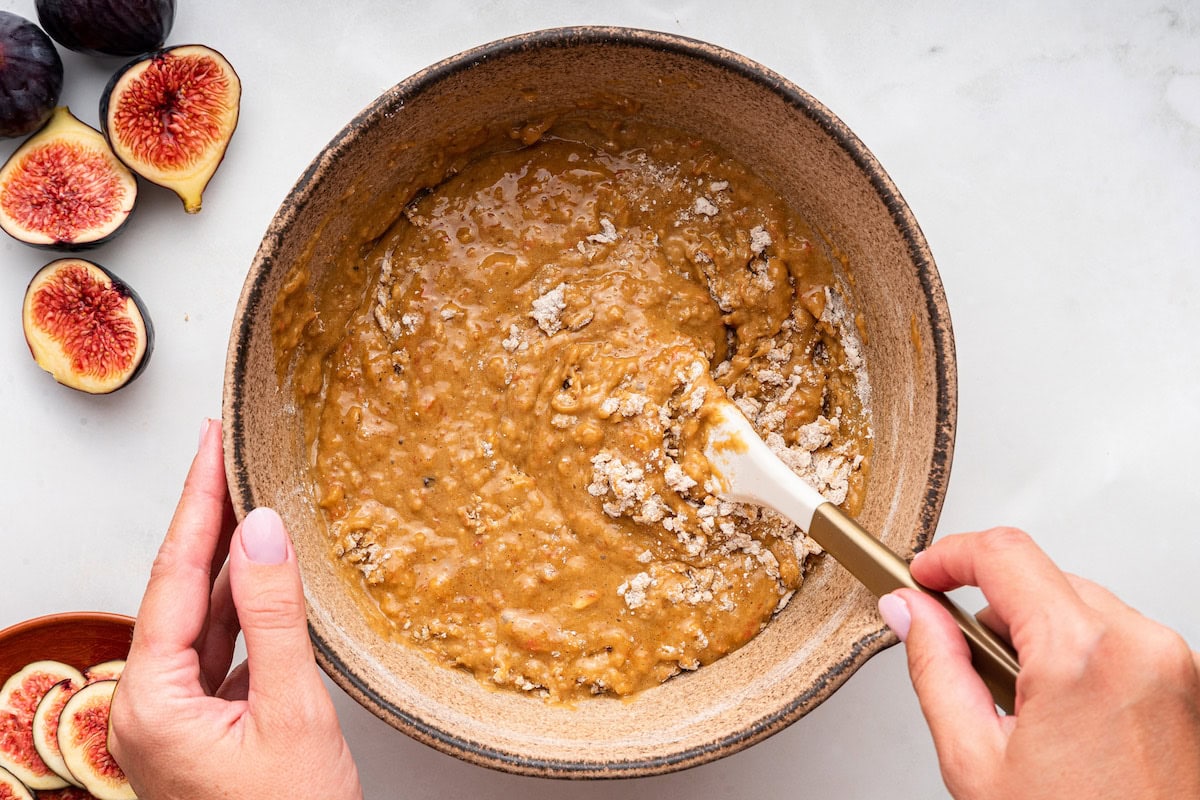 A womans hand uses a silicone spatula to combine the wet and dry ingredients for fig bread.