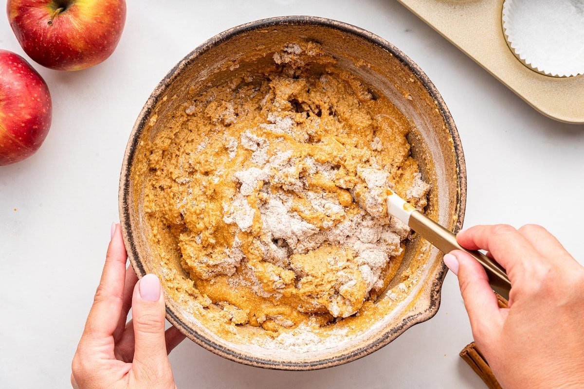 A woman's hand uses a silicone spatula to combine dry and wet ingredients used for apple muffins in a large mixing bowl.
