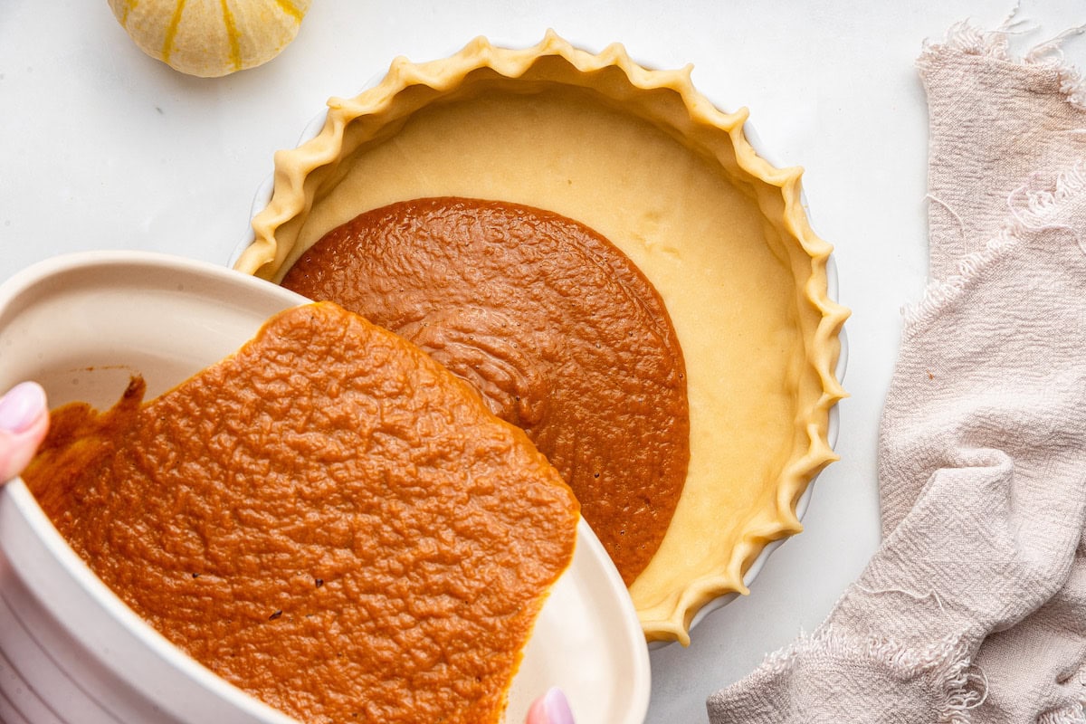 A woman's hands pours pumpkin pie batter into a prepared pie crust.