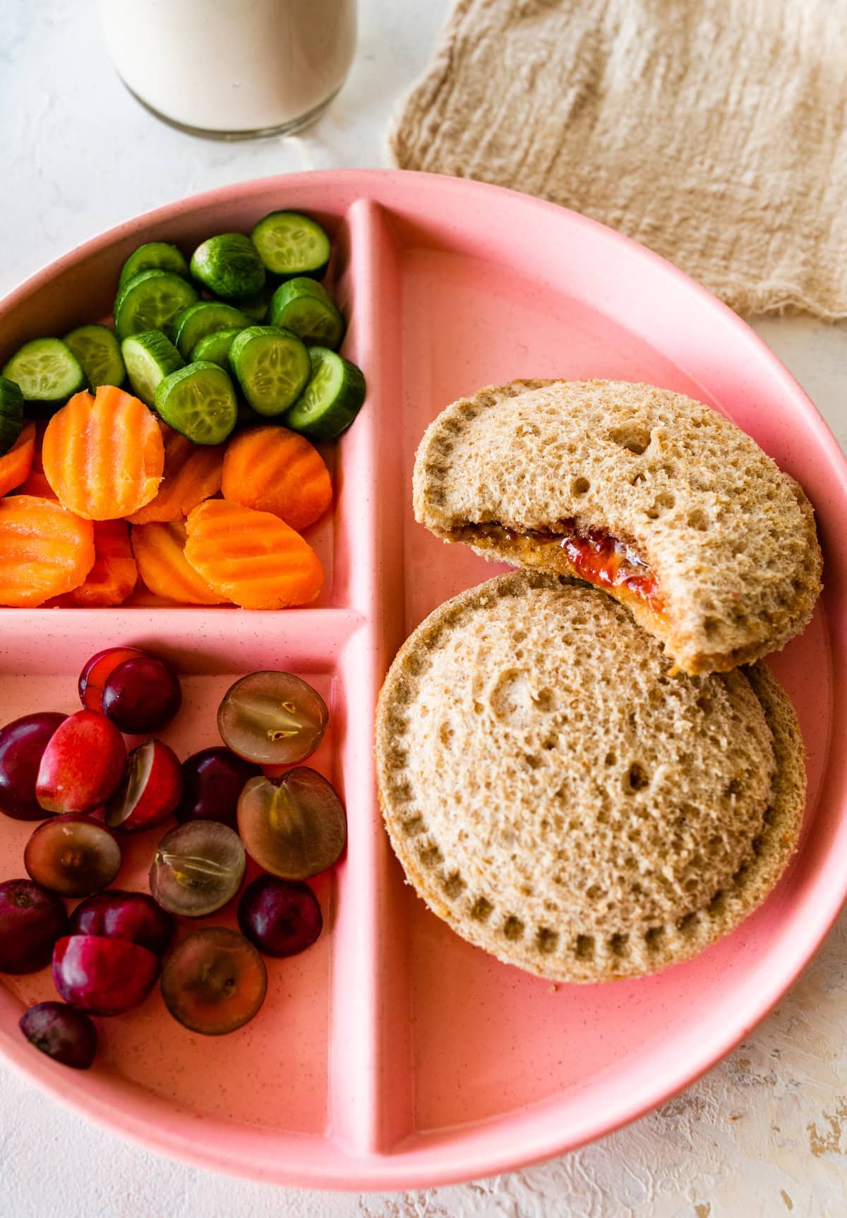 Two peanut butter and jelly uncrustables on a plate with grapes, carrots, and cucumbers on the side.