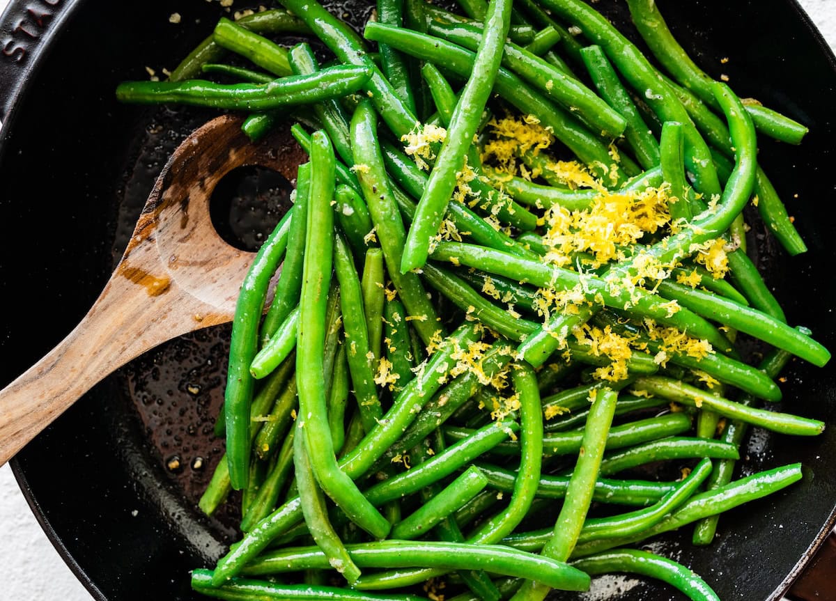 Sautéed green beans in a skillet with a wooden spoon topped with lemon zest.
