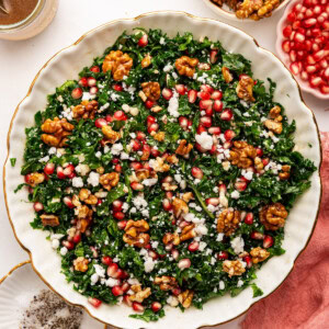 A kale salad topped with fresh pomegranate, goat cheese and candied walnuts. The salad is served in a white bowl.