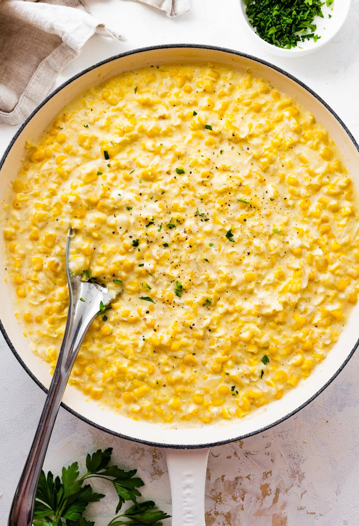 Creamed corn served in a white skillet with a silver spoon topped with freshly cracked pepper and fresh parsley.