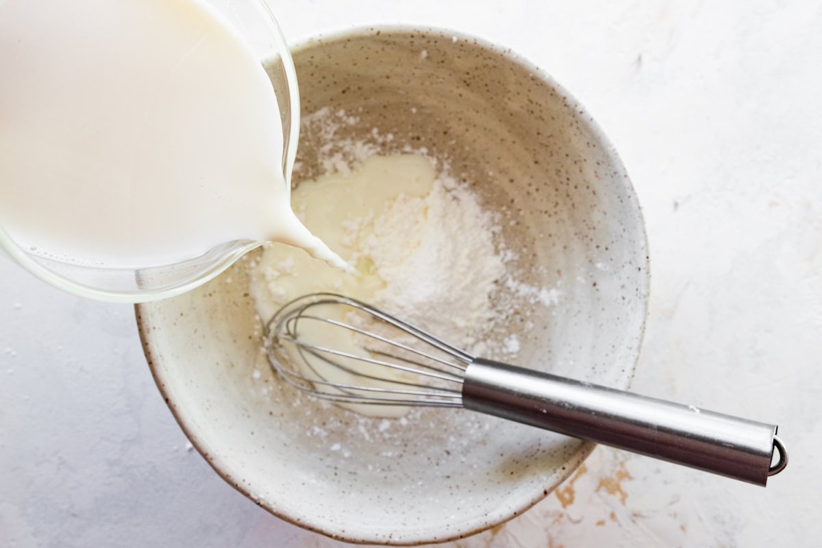 Milk being poured into a small speckled bowl with arrowroot powder. A whisk is in the bowl ready to mix it up.