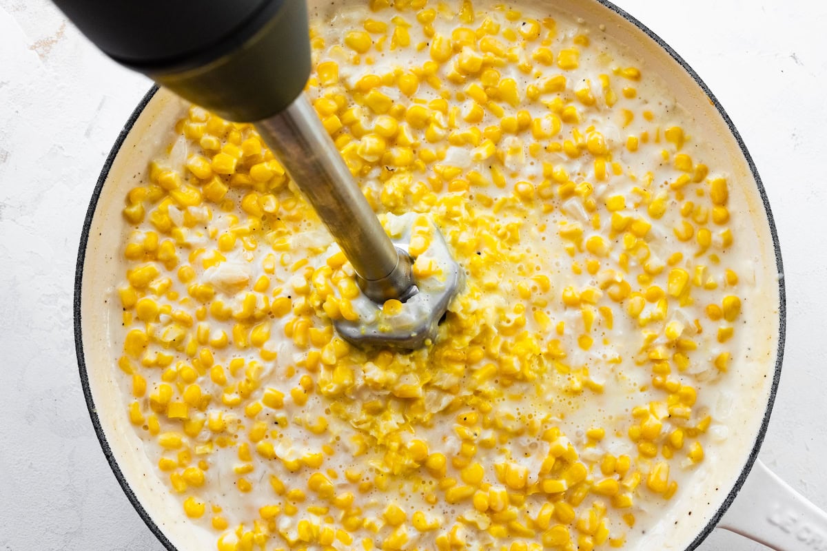 An immersion blender pureeing some of the creamed corn mixture in a white skillet.