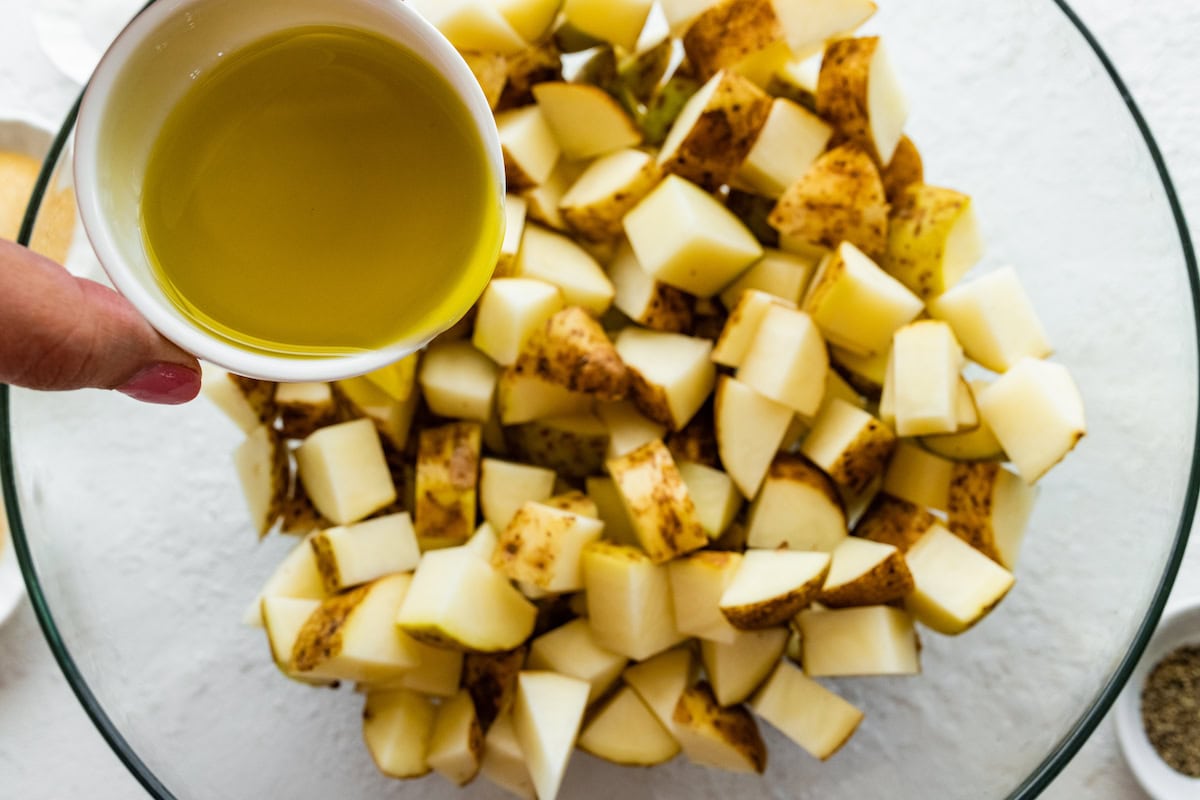 A woman's hand pours oil into a large glass bowl of diced potatoes.