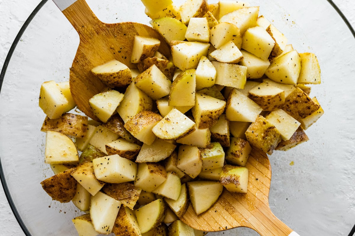 Diced potatoes seasoned in a large glass bowl with two serving spatulas.