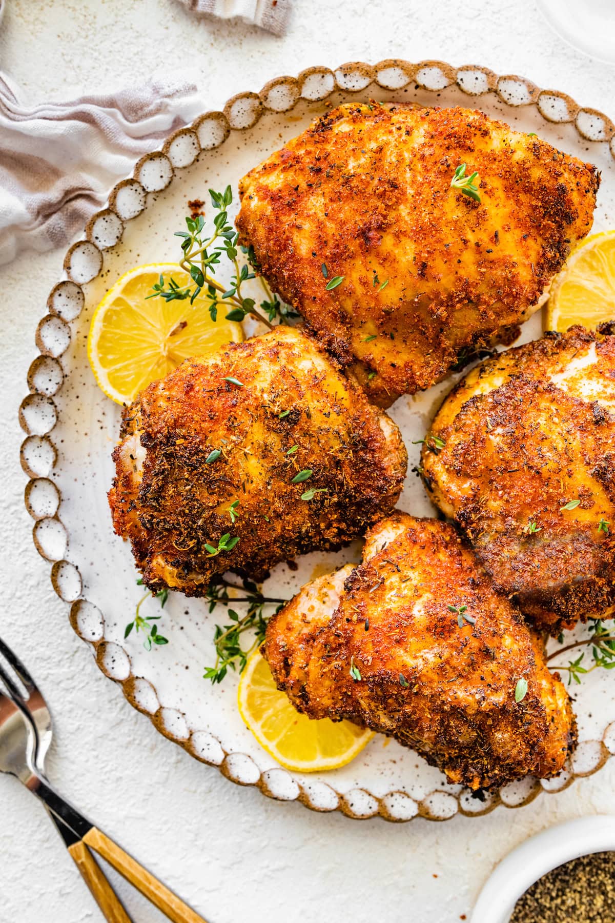 Four bone-in chicken thighs served on a white dinner plate topped with fresh parsley and thyme served with lemon slices.