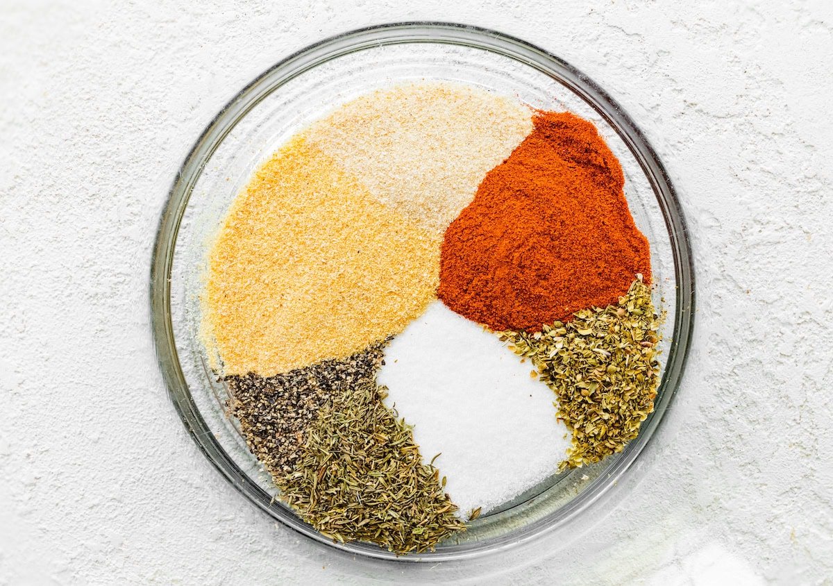 A blend of seasonings in a glass mixing bowl: garlic powder, onion powder, paprika, oregano, thyme, salt and pepper.