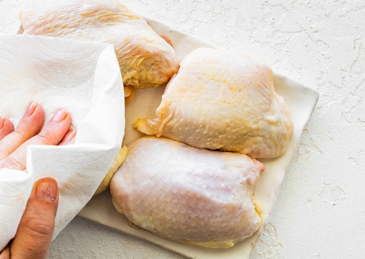 A woman's hand patting four bone-in chicken thighs dry with a paper towel.