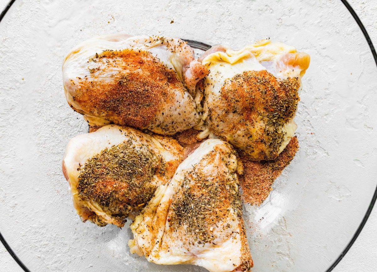 Four raw bone-in chicken thighs in a glass bowl tossed with seasonings.