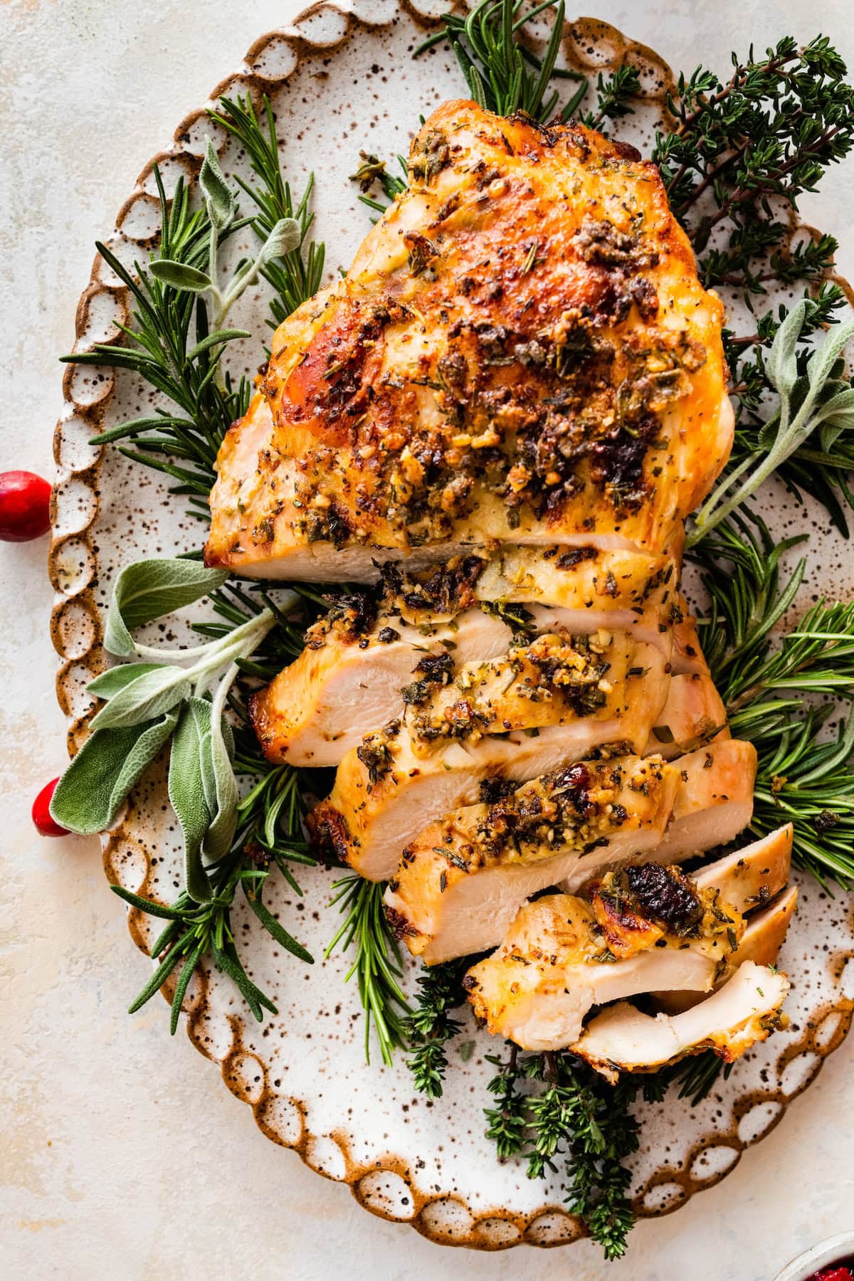 An air fryer turkey breast sliced sitting on a bed of fresh herbs and cranberries on a white platter.
