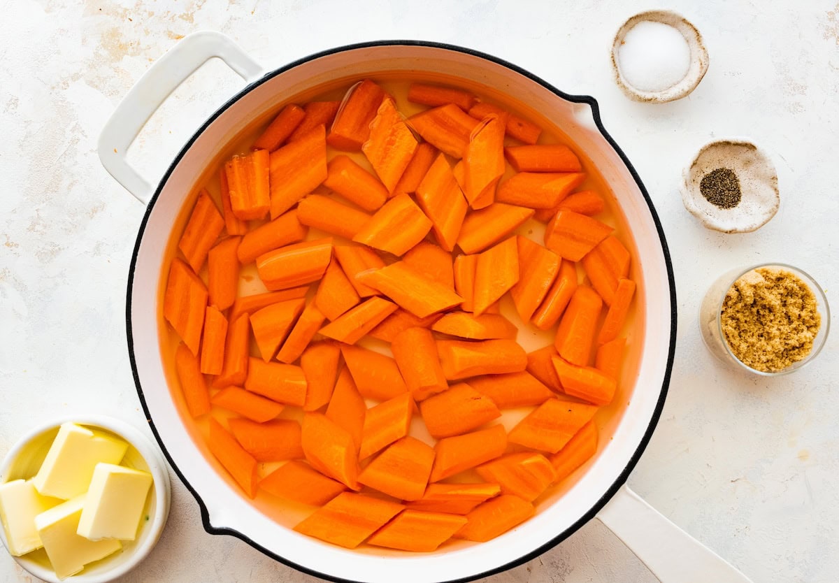Sliced carrots simmering in a pot of water. The pot is white and there are a few bowls around the pot filled with butter, salt, pepper and brown sugar.