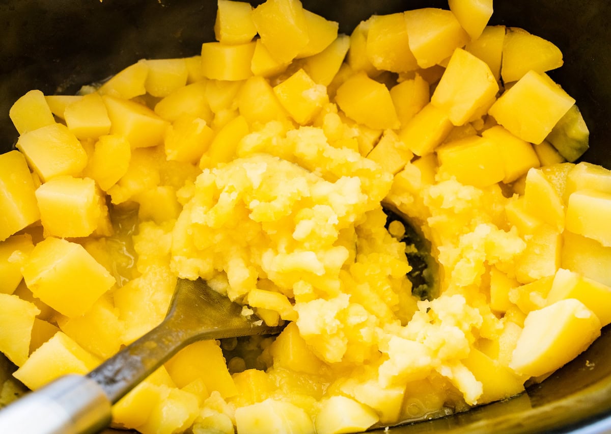 A potato masher mashing potatoes in a slow cooker.