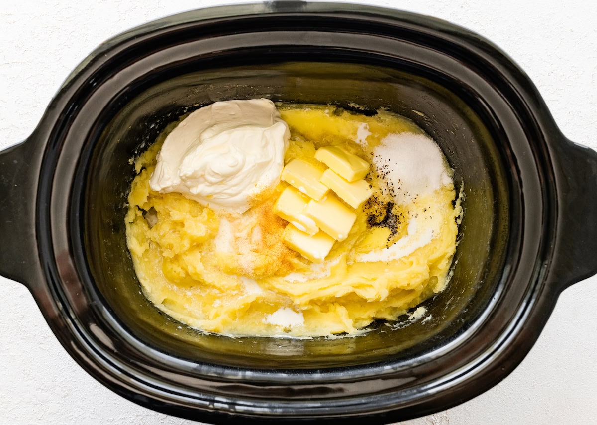 Mashed potatoes in a slow cooker with sliced butter, salt, pepper, sour cream, and milk.