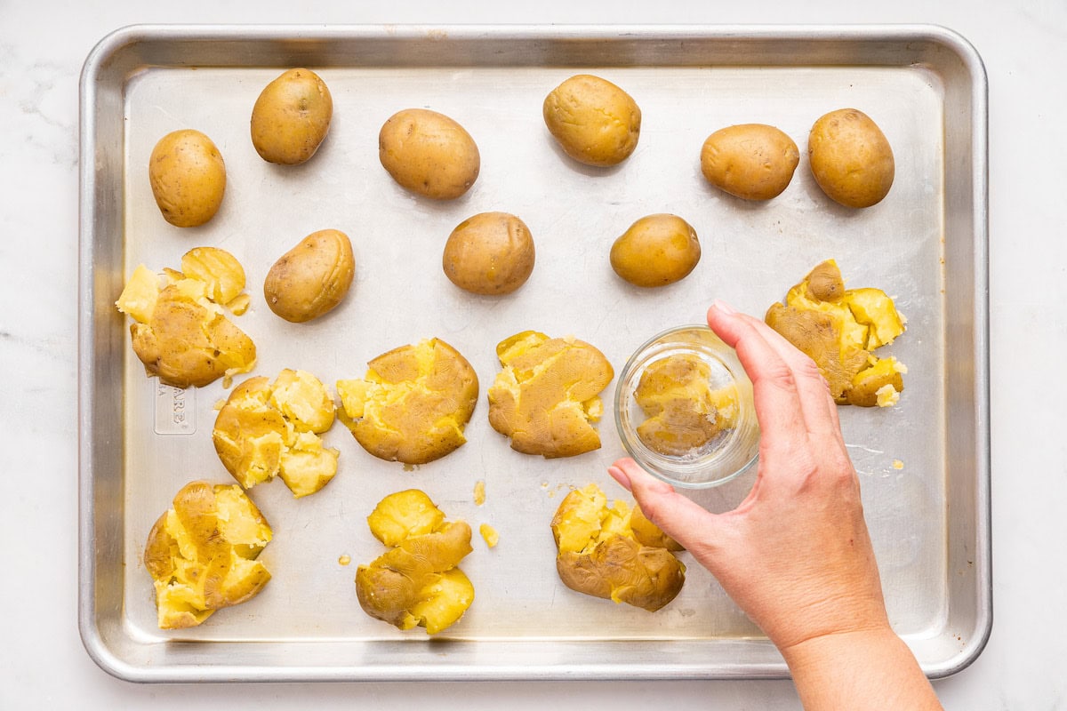 A womans hand uses a glass cup to smash cooked potatoes on a baking tray.
