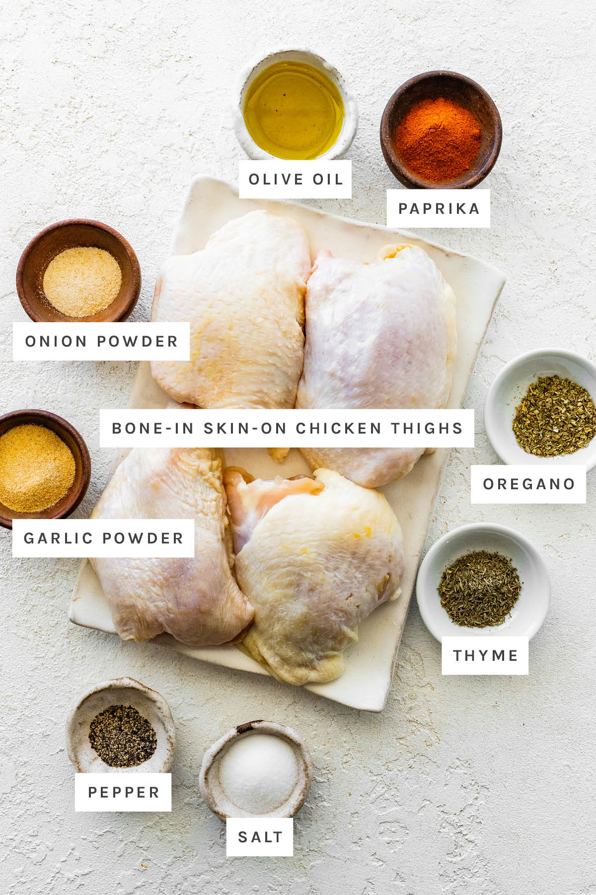 Ingredients measured out to make Air Fryer Chicken Thighs: olive oil, paprika, onion powder, garlic powder, bone-in skin-on chicken thighs, oregano, thyme, pepper and salt.