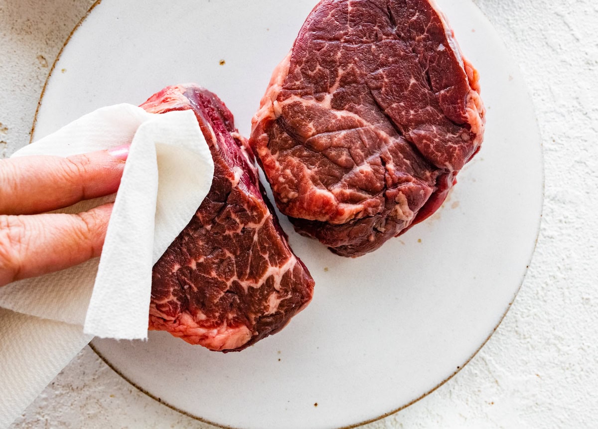 A woman's hand patting two filet mignons dry with a paper towel.