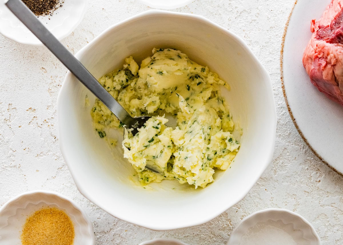 Mixed together garlic butter in a small white bowl. The butter is being mixed together with a silver spoon.