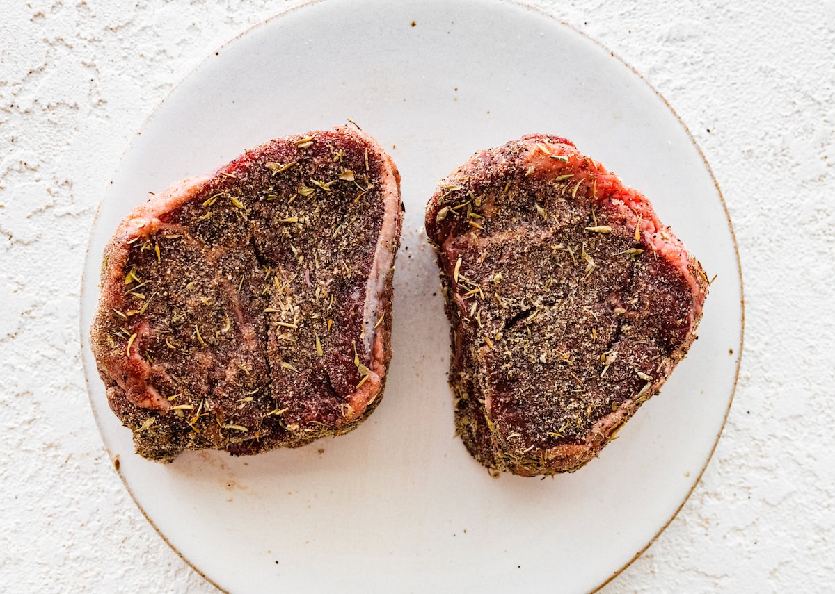 Two raw filet mignon pieces seasoned on a white plate.