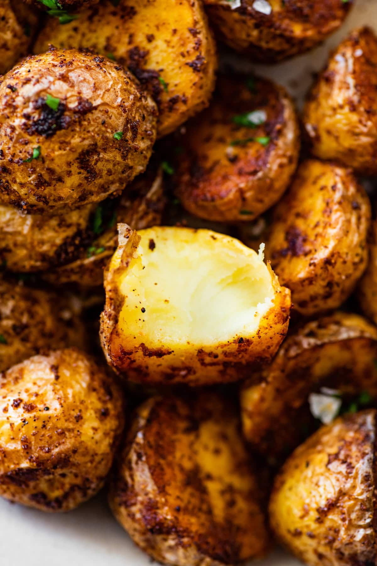 Crispy air fryer potatoes on a white plate garnished with fresh herbs and flaky sea salt. One potato has a bite taken from it.