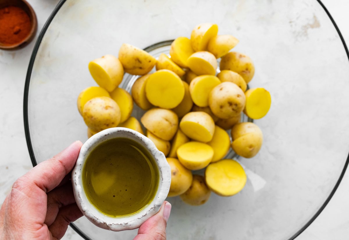 A womans hand holds a small container of oil over a large glass bowl of potato chunks.