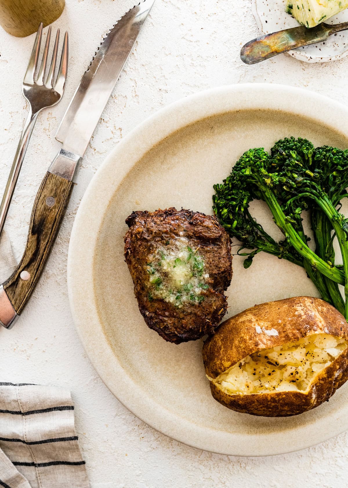A filet mignon served on a plate alongside broccolini and a baked potato. The filet is topped with garlic butter.