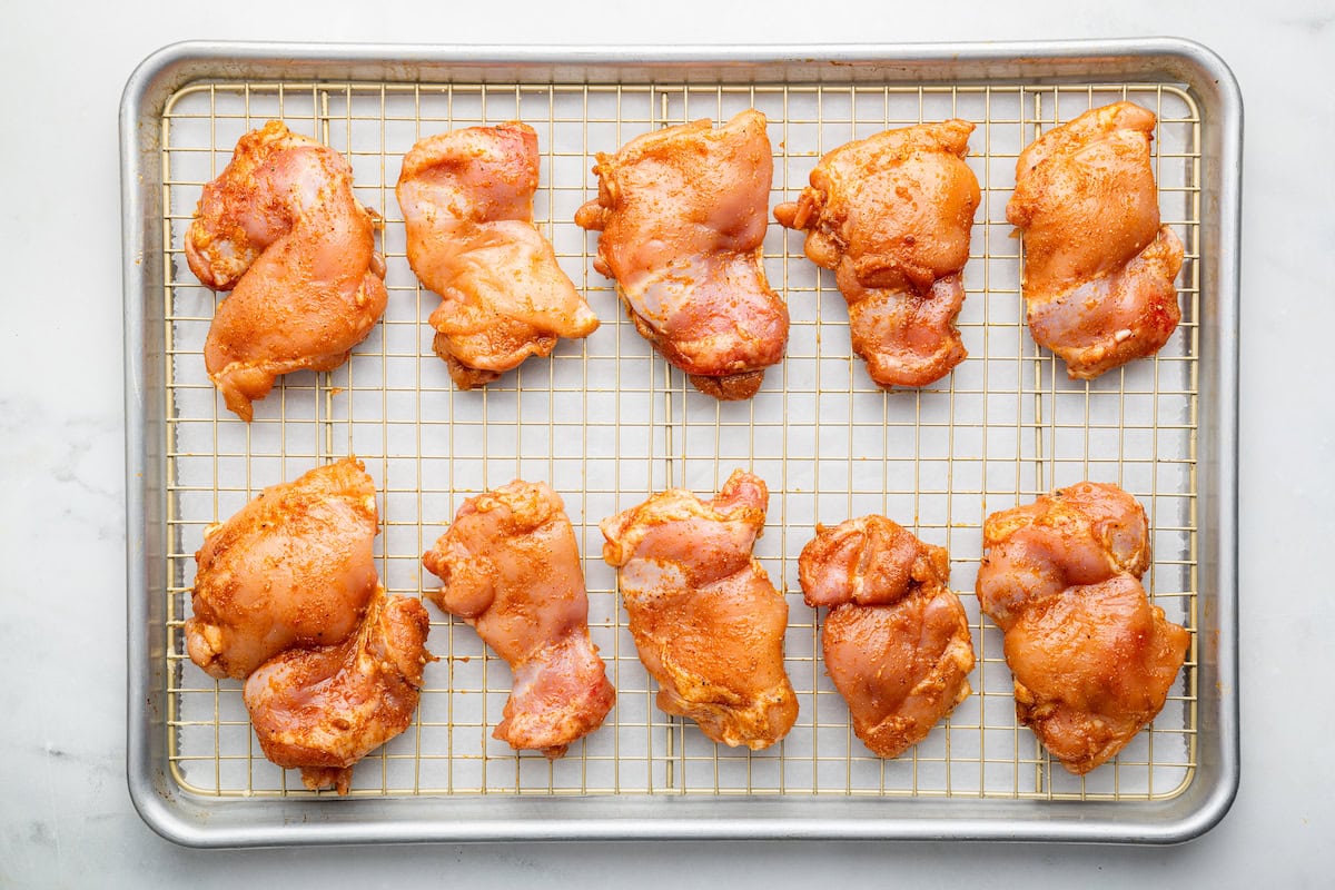 Raw and seasoned boneless chicken thighs on a metal rack on a baking tray.