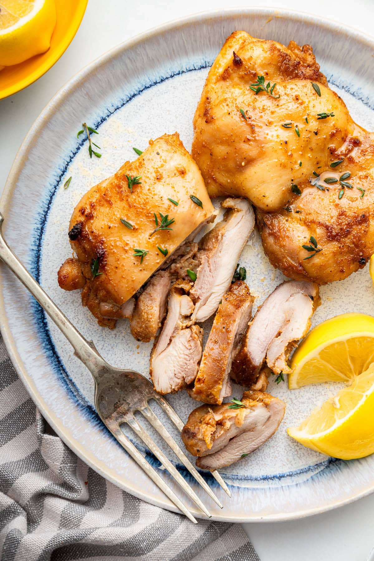 Two baked boneless chicken thighs on a plate with a lemon wedge and fork on the side. One chicken thigh is cut into bite size pieces.