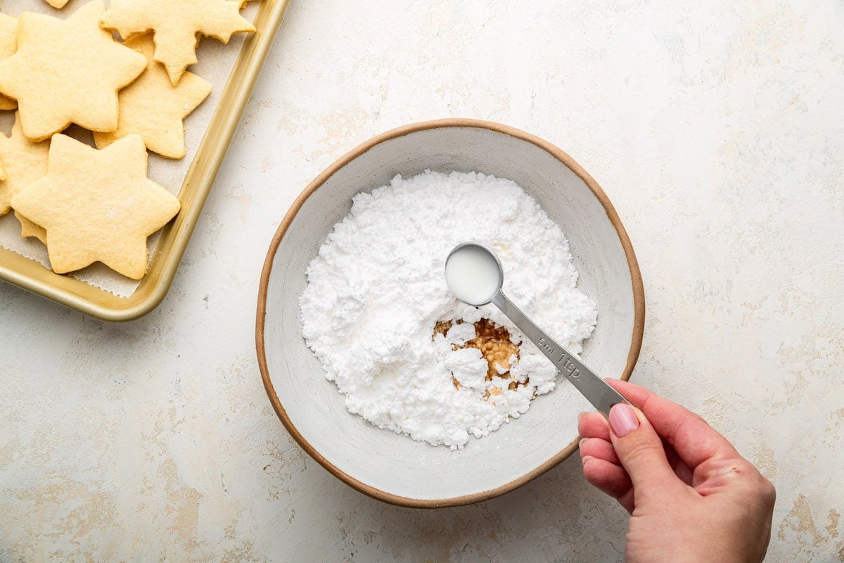 A woman's hand pours a teaspoon of milk in a bowl of powdered sugar to create an icing.
