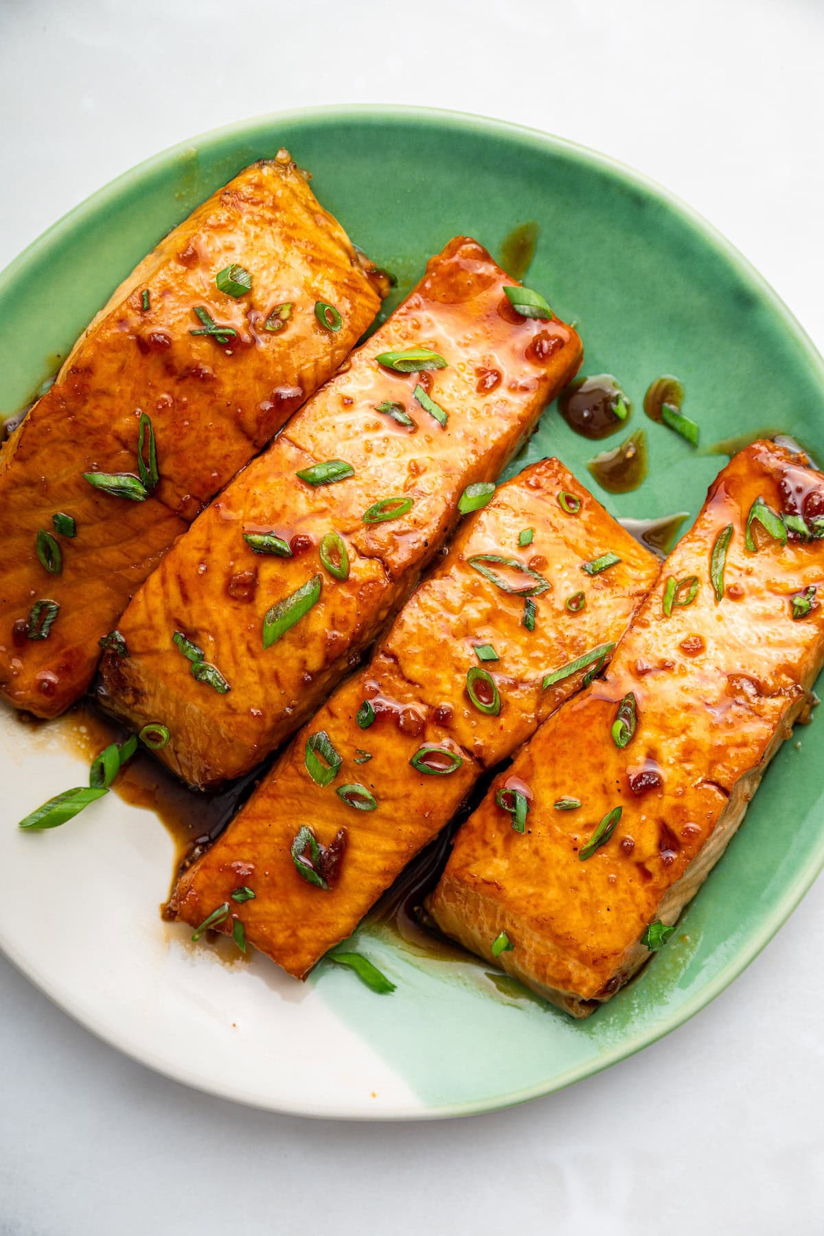 Four honey glazed salmon filets garnished with fresh chives on a green and white plate.