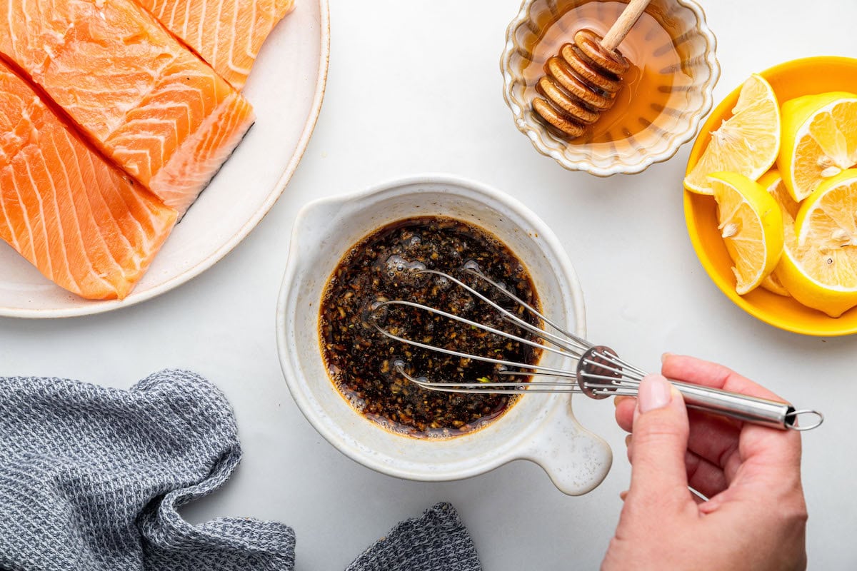 A woman's hand uses a metal whisk to whisk a honey based sauce in a small bowl.