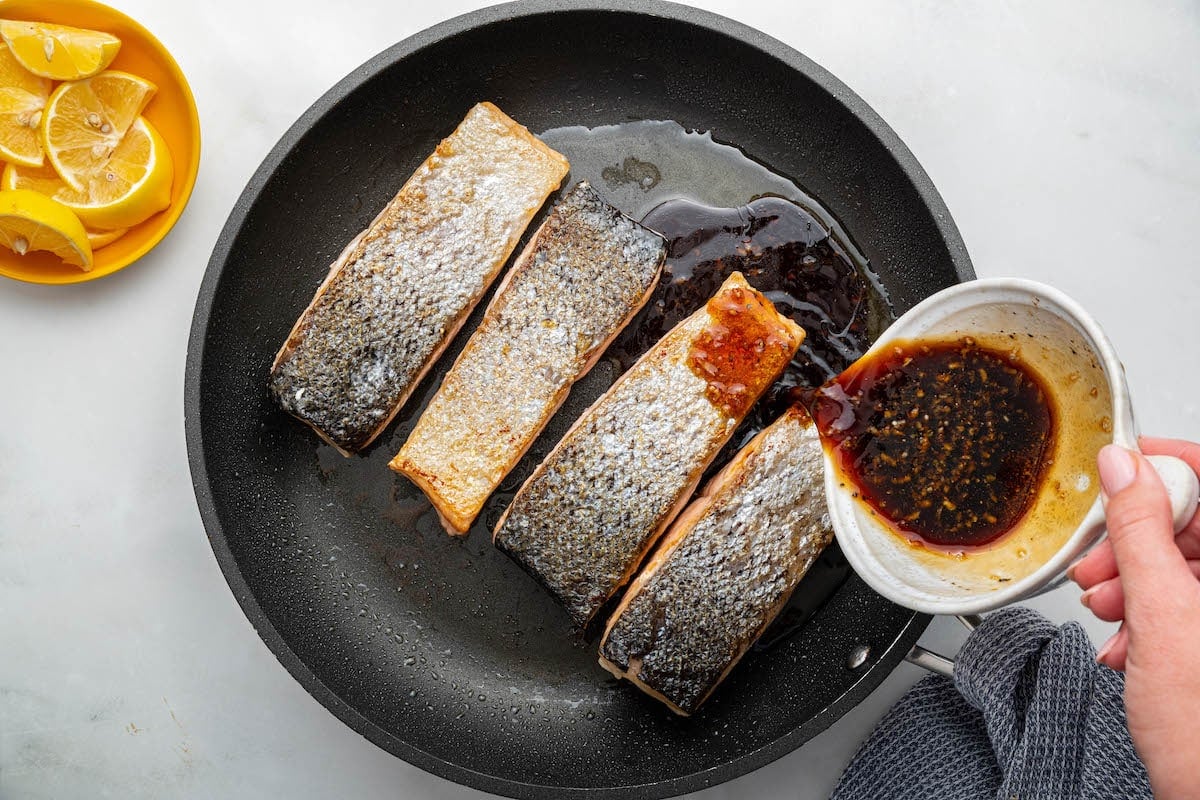 A woman's hand pours a honey based sauce over four salmon filets that are cooking in a skillet.