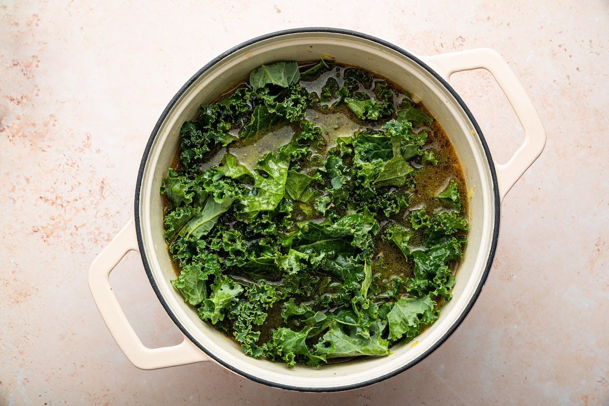Chopped kale being added to the sausage and kale soup in a pot.