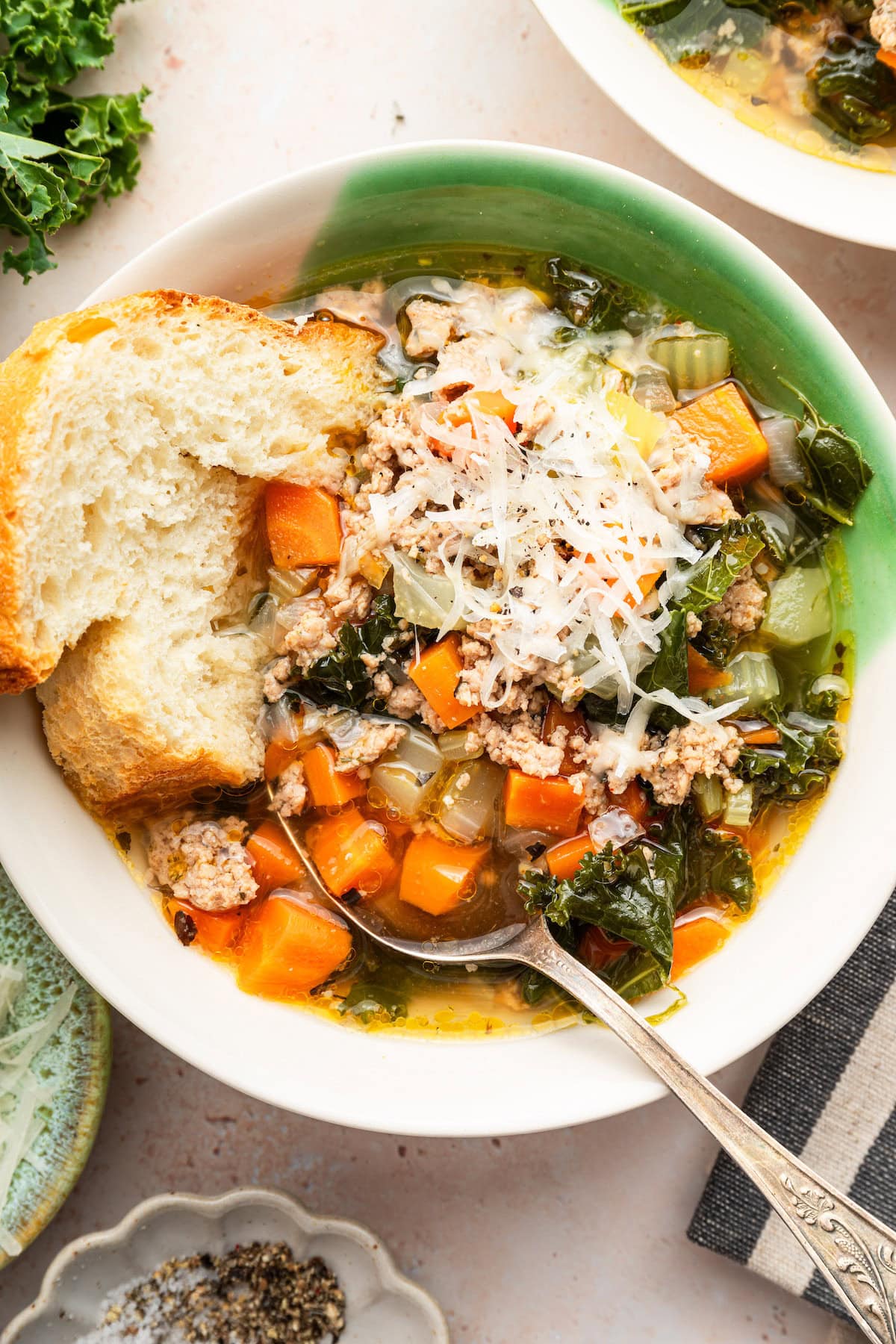 Sausage and kale soup served up in a green and white soup bowl. The soup is topped with grated parmesan cheese and served with a slice of bread and metal spoon.