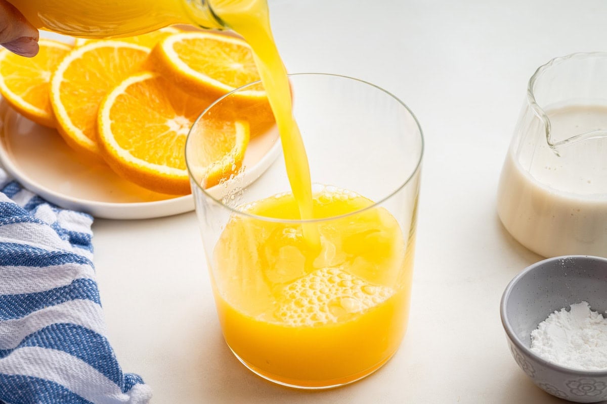 Orange juice being poured into a glass. Orange slices on a plate are in the background.
