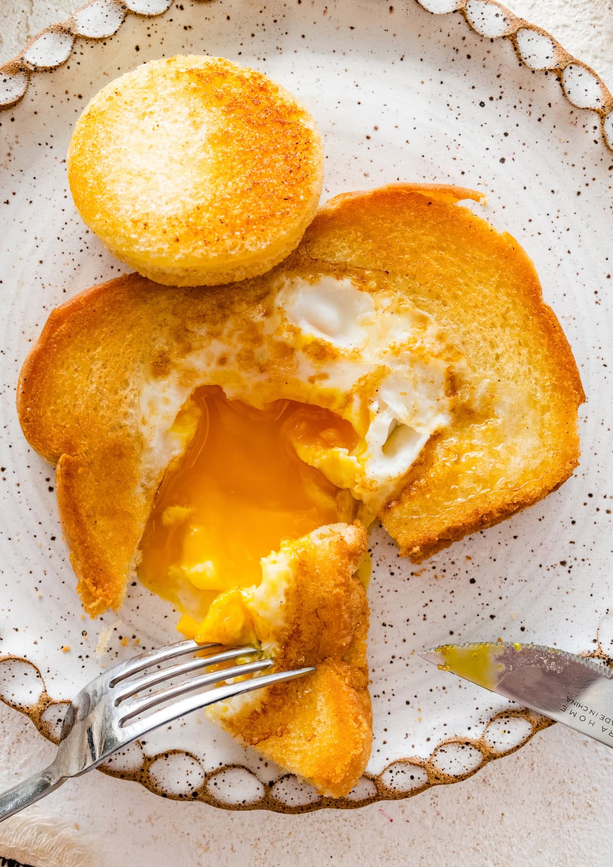 A slice of toast on a plate with a cooked egg in the center of it. A fork has removed a piece of the bread and the egg yolk is runny.