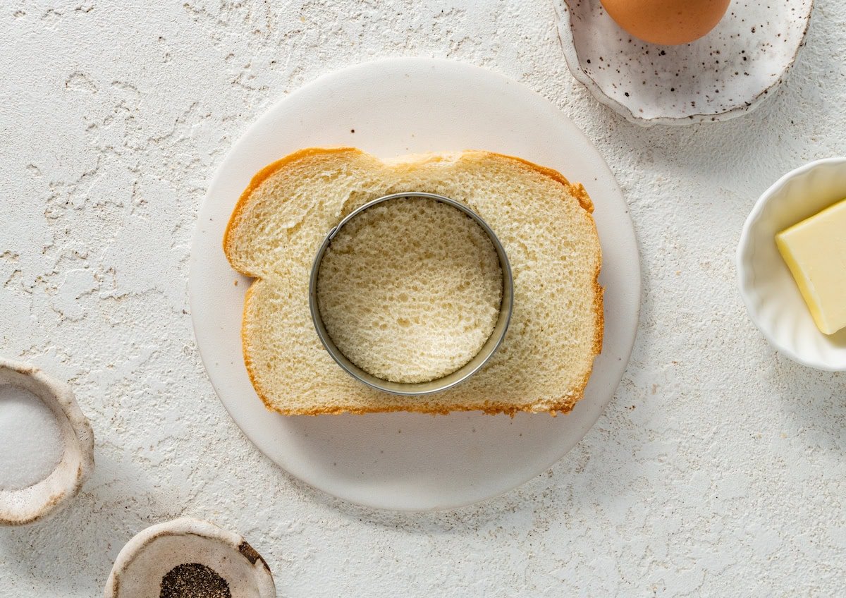 A round cookie cutter cutting out the middle of a slice of bread. The bread is on a white plate.