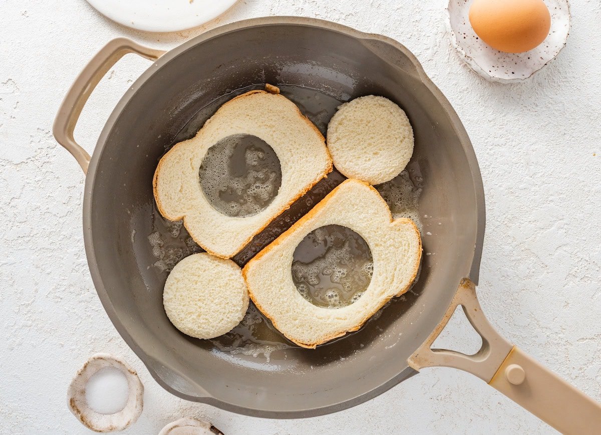 Two pieces of bread toasting in a skillet with melted butter. The center of the bread has been removed from both of the slices.