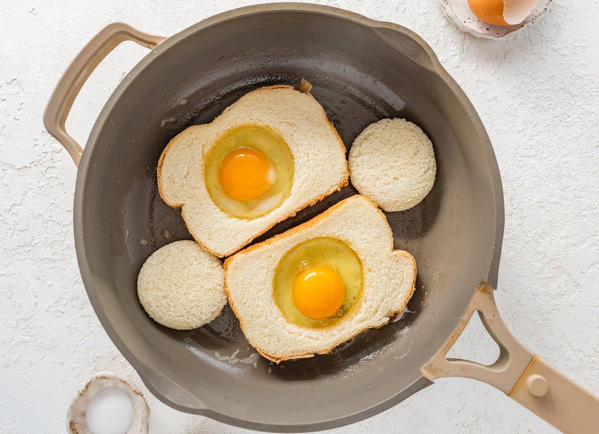 Two eggs cracked into the center of the two pieces of bread that are cooking on a skillet.