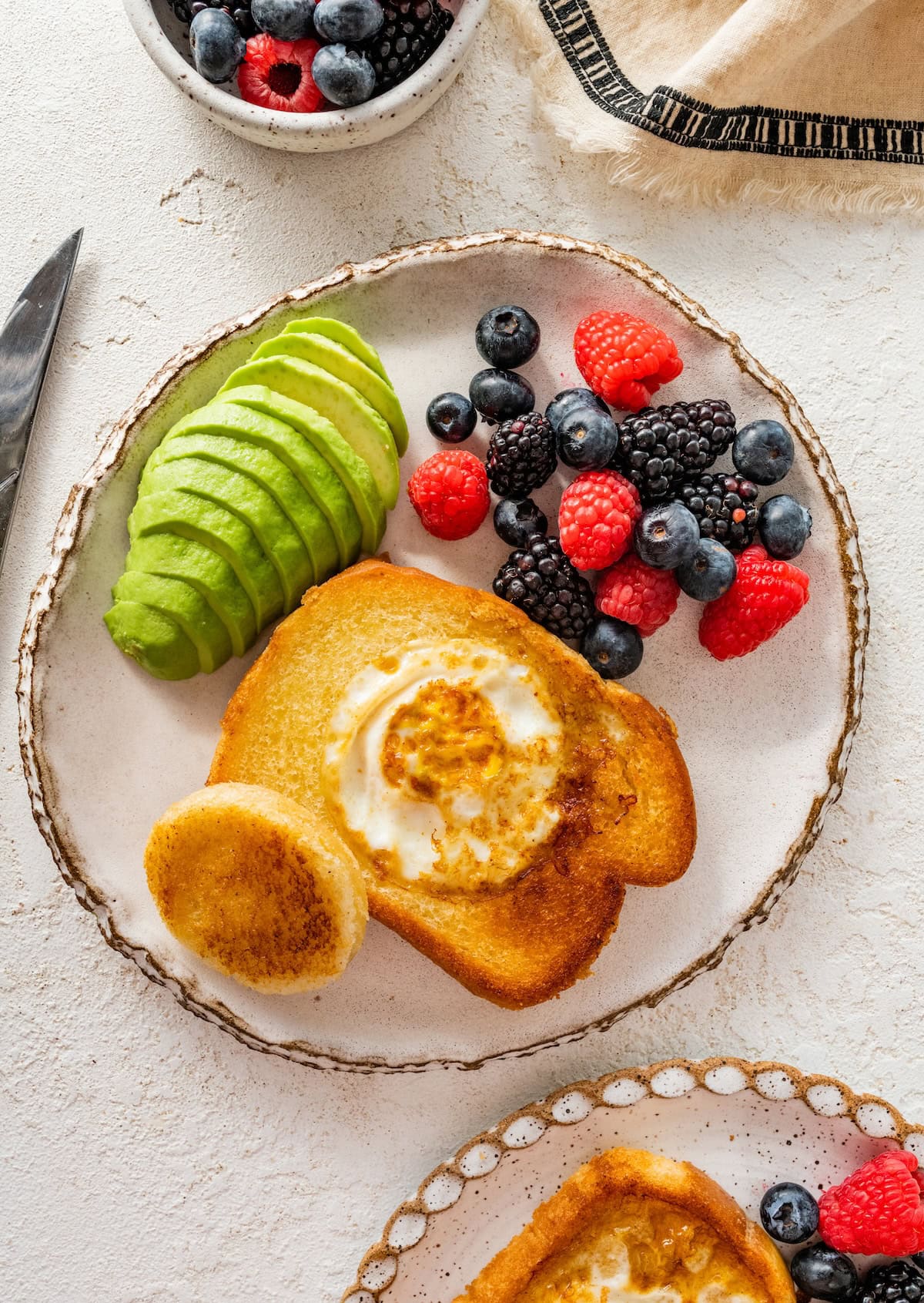 An egg in a hole served on a plate with fresh berries (raspberries, blueberries and blackberries) and avocado slices.