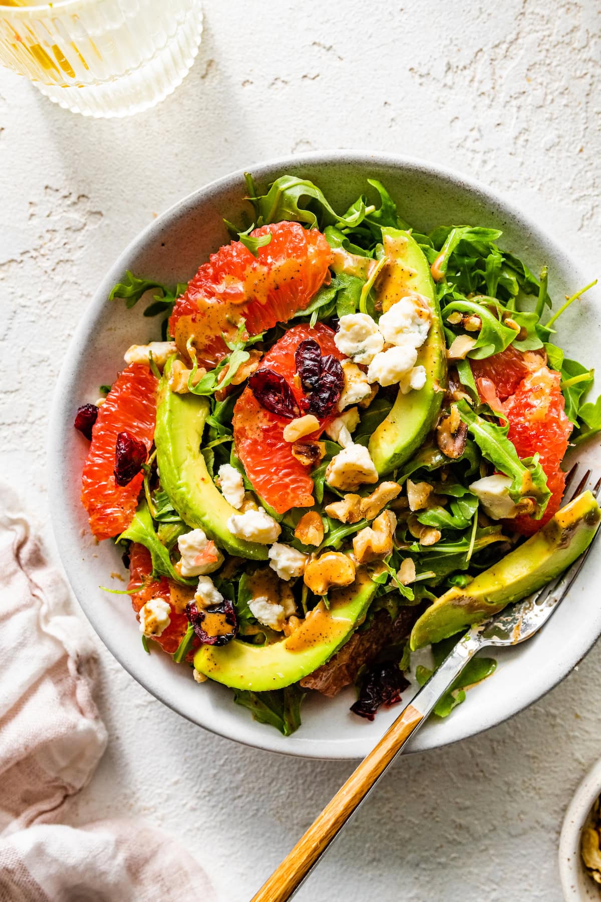 A salad with arugula, avocado, blue cheese, grapefruit, nuts, and cranberries in a white bowl with a fork.