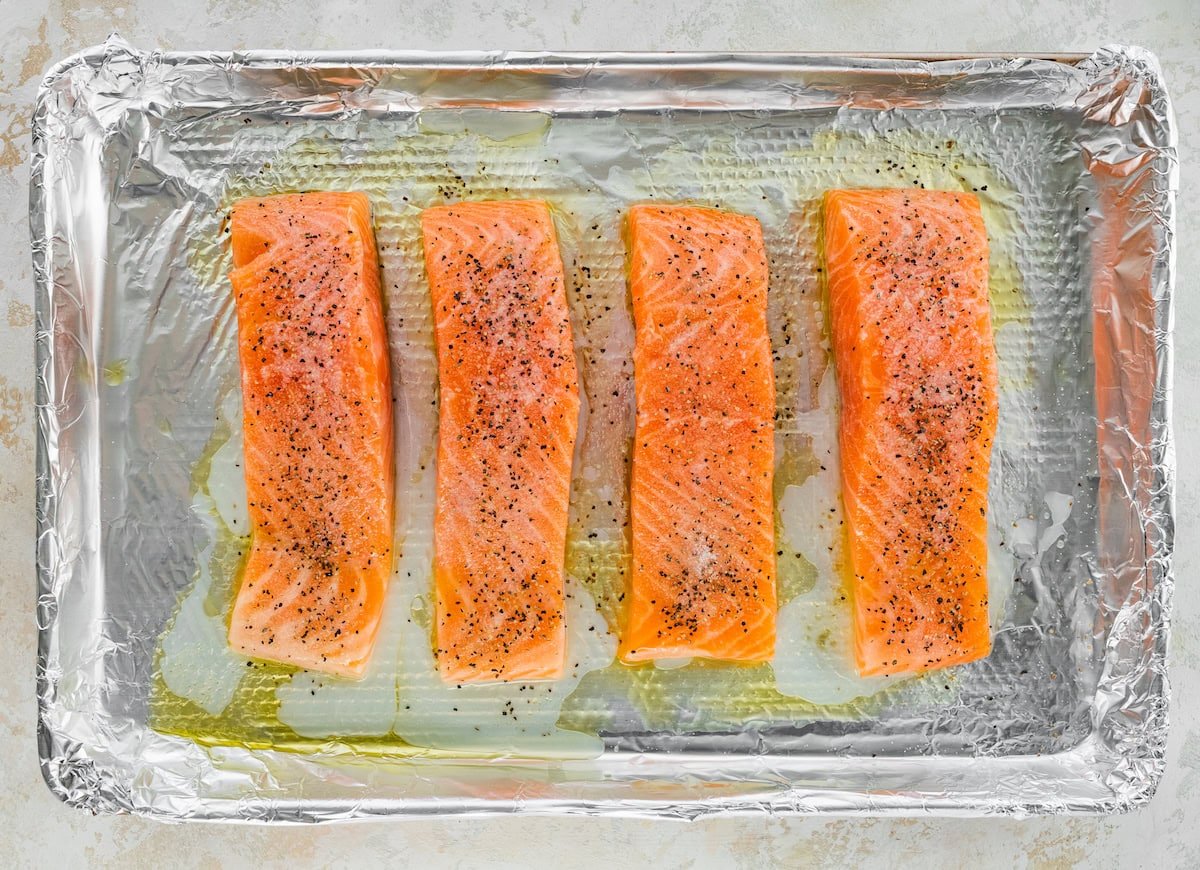Four raw salmon filets on a baking sheet lined with tin foil. The salmon is coated in olive oil, lemon juice, salt and pepper.