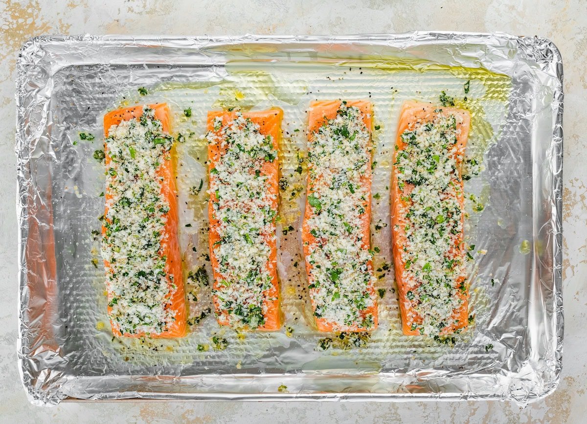 Four raw salmon filets on a baking sheet lined with tin foil. The salmon filets are topped with the parmesan herb mixture.