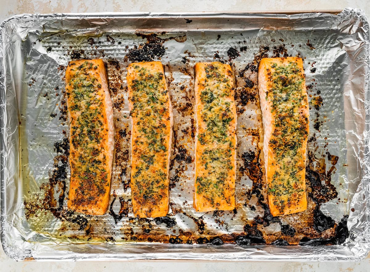 Four baked salmon filets on a baking sheet lined with tin foil. The salmon filets are topped with the parmesan herb mixture.
