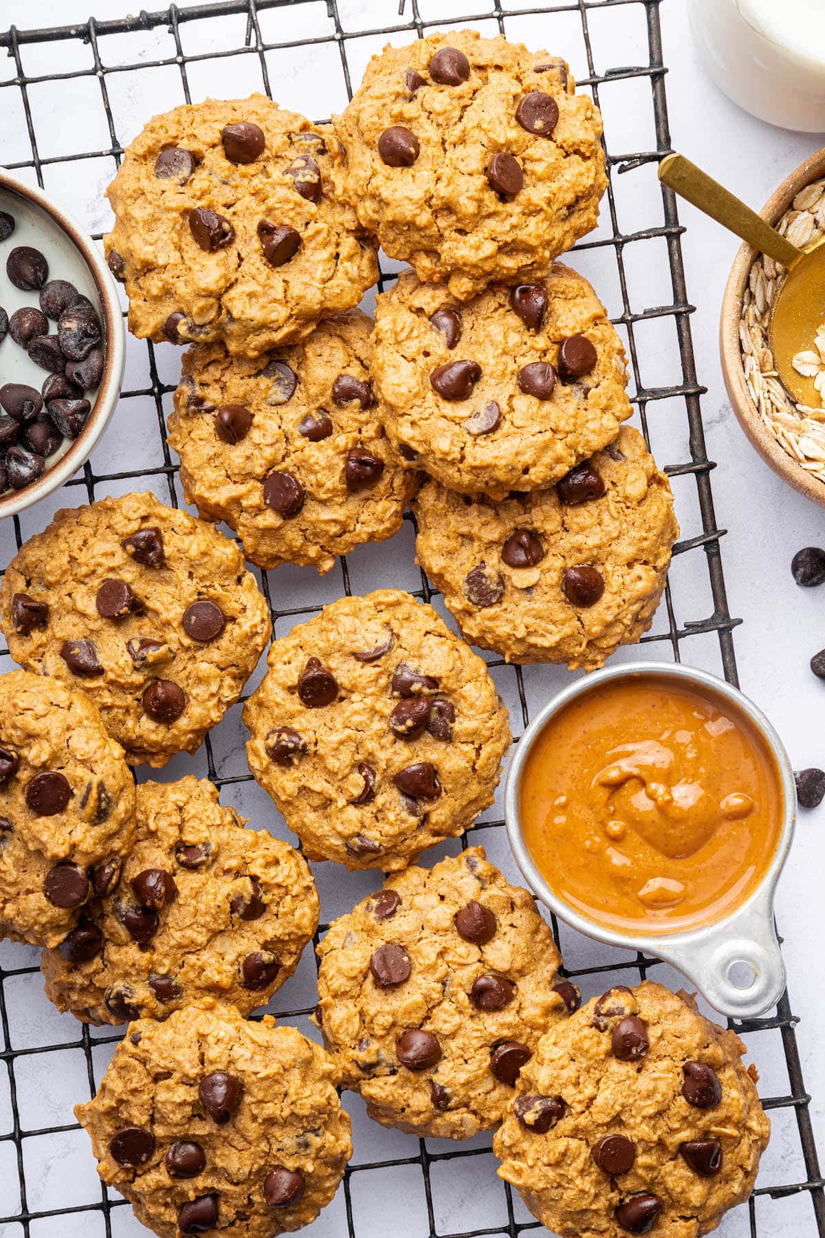 Multiple peanut butter oatmeal cookies with chocolate chips on a cooling rack near a small container of peanut butter.