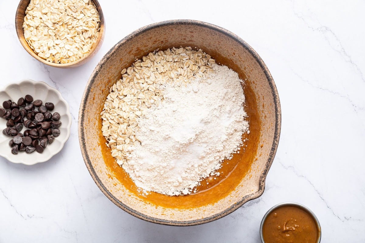 Dry ingredients including flour and oats added to a large mixing bowl of wet ingredients used for peanut butter oatmeal cookies.
