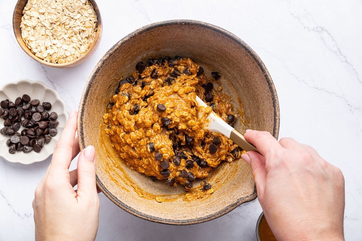 A womans hands using a silicone spatula to mix peanut butter oatmeal cookie dough in a large bowl.